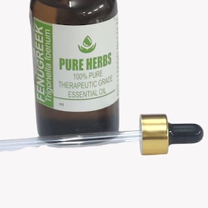 Pode incluir: Um frasco de vidro castanho com tampa branca e um rótulo com a inscrição FENUGREEK Trigonella foenum PURE HERBS 100% PURE THERAPEUTIC GRADE ESSENTIAL OIL. Um conta-gotas de vidro com colar dourado e bulbo preto está posicionado à frente.