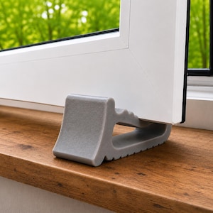 May include: A grey, wedge-shaped door or window stopper with a textured surface and a stepped, notched design on the top edge. The object is positioned underneath the bottom corner of a white window frame to hold it in an open position.
