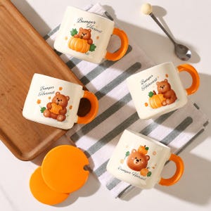 Könnte beinhalten: Vier cremefarbene Keramiktassen mit orangefarbenen Henkeln, jeweils mit einer braunen Bärenillustration, einem Kürbis und dem Text Bumper Harvest. Zwei orangefarbene runde Deckel und ein silberner Löffel mit einem kleinen Holzgriff.