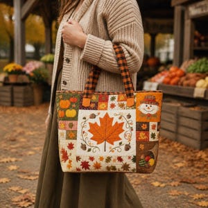 May include: A quilted tote bag featuring a patchwork design with autumn motifs including a large central maple leaf, pumpkins, acorns, a scarecrow face, and a cornucopia. The bag has two black and orange buffalo plaid fabric handles.