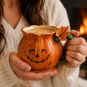 Peut inclure: Un mug en céramique en forme de citrouille avec une finition orange brillante, présentant un visage de citrouille sculpté souriant, un bord festonné et une anse verte texturée ornée d'une petite feuille d'érable brune, rempli d'une boisson crémeuse.
