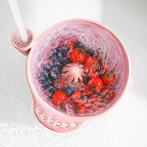 Könnte beinhalten: Ein rosafarbenes Keramiksieb mit strukturierter Oberfläche, einem zentralen Abflussdom und einem seitlichen Griff mit fünf kreisförmigen Löchern. Die Schale ist mit frischen Blaubeeren und roten Kirschtomaten gefüllt, die unter Wasser abgespült werden.