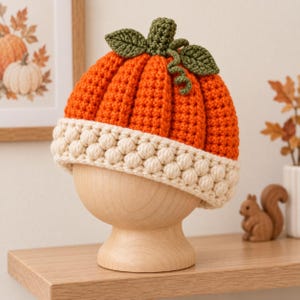 May include: A crochet hat shaped like a pumpkin, featuring a vibrant orange ribbed body, a cream-colored brim with a textured bobble stitch pattern, and a green stem with two small leaves and a curly vine detail on the very top of the crown.