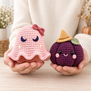 May include: A light pink crocheted ghost figure with a small bow, dark blue crescent moon eyes, and embroidered stars, alongside a dark purple crocheted pumpkin figure wearing a tan pointed hat with a green leaf and pink cheeks.