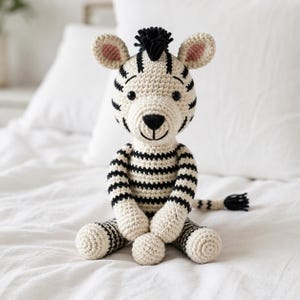 Può includere: Un peluche a forma di zebra realizzato all'uncinetto con un corpo color crema a strisce nere orizzontali, una criniera in filo nero e piccoli accenti rosa all'interno delle orecchie. Il giocattolo ha occhi ricamati neri, naso nero e una lunga coda a strisce.