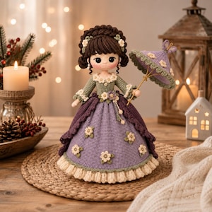 May include: A handcrafted crochet doll with dark brown hair styled in a braided updo, wearing a long dress in shades of lavender and sage green with small white flower appliqués and a cream-colored ruffled hem, holding a matching lavender parasol.