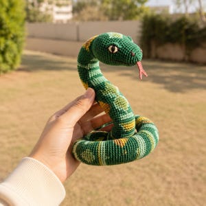 May include: A crocheted snake toy featuring a green and yellow patterned body with a textured surface. The snake has a coiled base, a raised head, and a small red forked tongue protruding from its mouth. The eyes are dark with light accents.