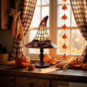 May include: A stained glass lamp shaped like a witch hat with orange and purple panels, resting on a dark metal base. The scene includes several carved jack-o'-lanterns, ears of dried corn, and a vertical string of small orange pumpkin ornaments.