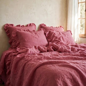 Pode incluir: Um conjunto de roupa de cama em rosa seco, composto por uma capa de edredão e quatro fronhas a condizer. Cada peça é adornada com um rebordo de folhos. O tecido tem uma textura visível e o conjunto apresenta uma cor rosa uniforme.