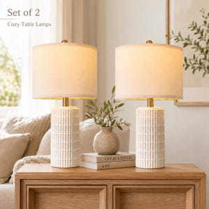 May include: A set of two table lamps featuring cylindrical white bases with a vertical grid texture and gold-toned accents. Each lamp is topped with a drum-shaped off-white fabric shade. The text Set of 2 Cozy Table Lamps is visible.
