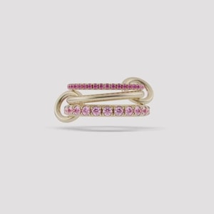 May include: A gold ring featuring three interconnected bands. The top and bottom bands are set with a row of small, round, pink gemstones, while the middle band is a smooth, plain gold hoop. The design creates a layered, linked appearance.
