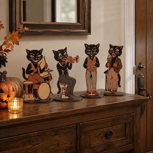May include: Four freestanding black cat figures arranged in a jazz band formation. One cat plays drums, another plays a trumpet, the third plays a clarinet, and the fourth sings into a microphone. Each cat wears vintage-style clothing on a flat base.
