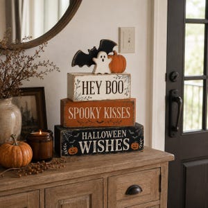 May include: A stack of three rectangular wooden blocks with Halloween themes. The top block is white with the text HEY BOO and a bat, ghost, and pumpkin topper. The middle block is orange with SPOOKY KISSES. The bottom block is black with HALLOWEEN WISHES.