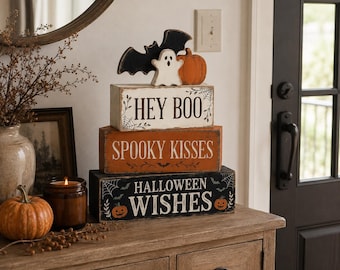 May include: A stack of three rectangular wooden blocks with Halloween themes. The top block is white with the text HEY BOO and a bat, ghost, and pumpkin topper. The middle block is orange with SPOOKY KISSES. The bottom block is black with HALLOWEEN WISHES.