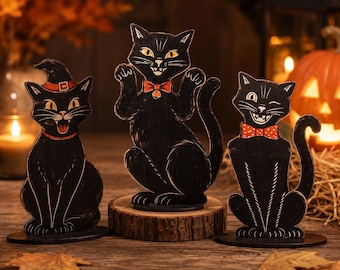May include: Three black cat figurines with yellow eyes and red accents. The left cat wears a witch hat, the centre cat has a red bow tie with a bell, and the right cat wears a red polka-dot bow tie. Each cat is mounted on a circular base.