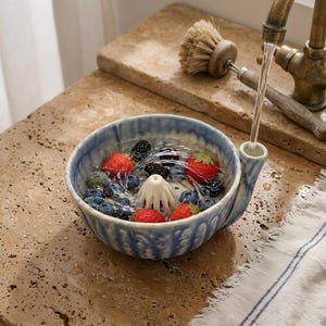 May include: A blue and white ceramic bowl with a built-in juicer cone in the centre, filled with water, fresh strawberries, and blueberries. A stream of water flows from a tap into the small spout attached to the side of the bowl.
