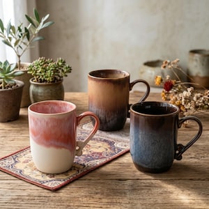 Op de afbeelding: Drie keramische mokken met reactieve glazuurafwerkingen. Eén mok heeft een verloop van koraalroze en crème, een andere een warme bruine en beige ombre, en de derde een mix van diepblauw en donkerbruin. Elke mok heeft een gebogen handvat en een ronde vorm.