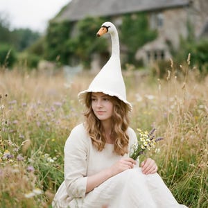 May include: A white felted hat shaped like a swan with a long neck extending upwards and a pointed orange beak at the top. The hat features a wide, slightly ruffled brim that sits around the head, crafted from a soft, off-white material.