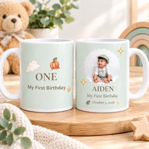 May include: Two white ceramic mugs with a light green background. The left mug features a pumpkin, a cloud, and the text ONE My First Birthday. The right mug displays a circular photo of a baby, the name AIDEN, My First Birthday, and October 5, 2026.