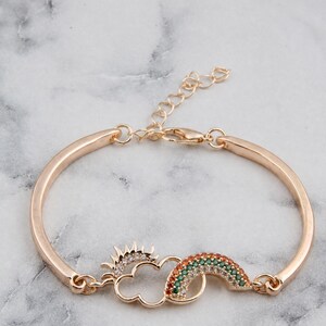 May include: A gold-toned metal bracelet featuring a central charm of a sun, a cloud, and a rainbow. The sun is accented with clear crystals, while the rainbow is inlaid with small orange, yellow, and green stones. It includes a lobster clasp closure.