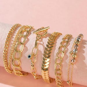 May include: A set of six gold-tone bracelets featuring various chain styles, including a curb chain, an oval link chain with a small blue evil eye charm, a toggle clasp chain, a chevron link chain, and a delicate chain with metallic beads.