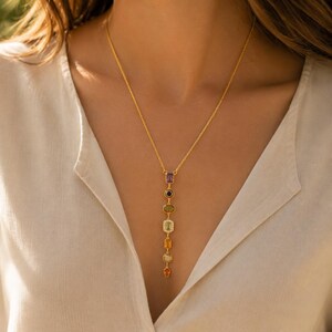 May include: A gold chain necklace featuring a vertical drop pendant composed of seven multi-colored gemstones in various shapes, including purple, dark blue, green, light green, yellow, clear, and orange stones set in gold metal settings.
