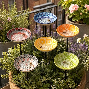 May include: A set of five decorative ceramic bowls on black metal stakes, each featuring a unique colourful pattern in blue, red, orange, yellow, and green. The bowls are filled with clear glass beads and are surrounded by green plants and bees.