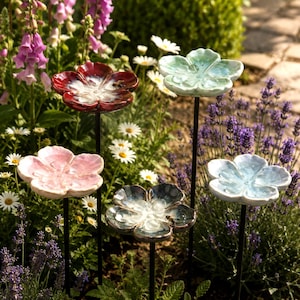 May include: Four ceramic flower-shaped garden stakes in red, light green, light pink, and light blue. Each flower features a glossy finish and is mounted on a thin black metal rod, designed to be placed in soil among garden plants and flowers.