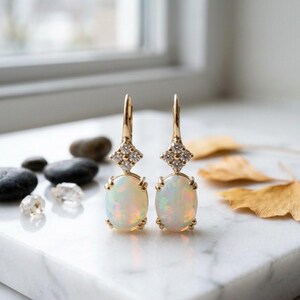 May include: A pair of gold drop earrings featuring oval-shaped opals suspended from a diamond-encrusted square setting. The earrings have a lever-back closure and display iridescent flashes of color within the white opal stones.