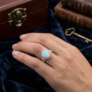 May include: A silver-toned ring featuring a large, oval-shaped iridescent opal center stone surrounded by a halo of small, clear round-cut stones. The band is split and encrusted with additional small, clear stones along the sides.