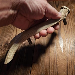 May include: A rustic cabinet pull made from a section of deer antler. The antler is a light tan color with a natural, textured surface. Two metal posts are attached for mounting. A unique accent for furniture.