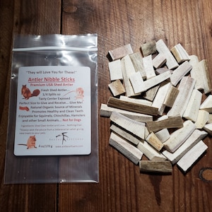 May include: A bag of antler nibble sticks for small animals. The bag is labeled "Antler Nibble Sticks" and "Premium USA Shed Antler". The antler nibble sticks are a natural organic source of minerals and promote healthy and clean teeth. The bag also states that the antler nibble sticks are not for dogs. The bag is on a wooden surface.