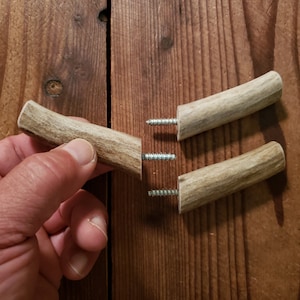May include: Three antler hooks with screws attached. The hooks are made of natural antler and are a light brown color. The screws are silver.