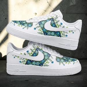 Peut inclure: Une paire de baskets blanches avec un motif floral composé de fleurs bleues et de feuilles vertes sur les panneaux latéraux et le bout. Les chaussures ont des lacets blancs, un logo Nike blanc sur le côté et le texte AIR gravé sur la semelle.