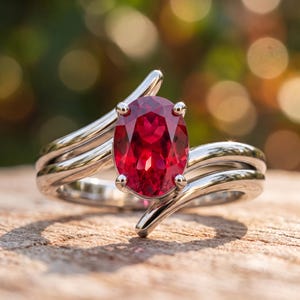 May include: A silver-toned ring featuring a large, oval-cut, deep red gemstone held in place by four prongs. The band consists of two smooth, polished metal strands that curve and overlap around the central stone in a bypass-style design.