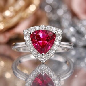 May include: A silver-toned ring featuring a large, triangular-cut vibrant red gemstone at its center. The red stone is surrounded by a halo of small, clear, round-cut white stones set into the metal frame, which extends into a smooth band.