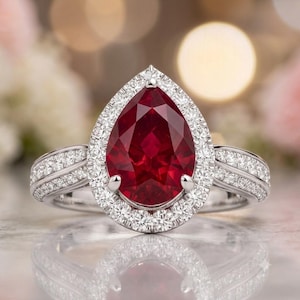 May include: A pear-shaped red gemstone ring set in a silver-toned metal band. The central red stone is surrounded by a halo of small white stones, with additional small white stones embedded along the split shank of the ring band.