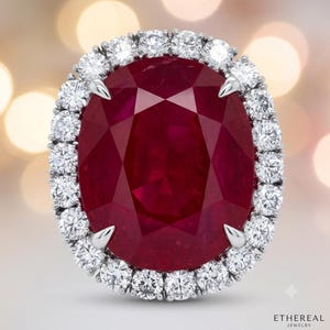 May include: A ring featuring a large, oval-cut deep red ruby centre stone held by four prongs. The ruby is surrounded by a halo of round, brilliant-cut white diamonds set in a metallic band. The text ETHEREAL JEWELRY is visible at the bottom.