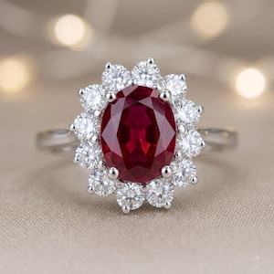 May include: A silver-toned ring featuring a large, oval-cut deep red gemstone at the centre, surrounded by a halo of twelve small, round, clear white stones arranged in a floral pattern, set on a polished metal band.