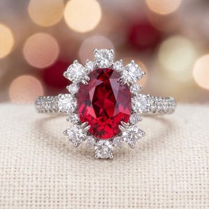 May include: A silver-toned ring featuring a large, oval-cut deep red gemstone at the center. The central stone is surrounded by a halo of small, round, clear white stones, with additional clear stones set along the band of the ring.