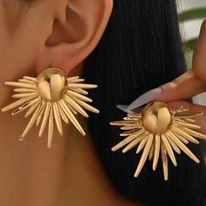 May include: A pair of gold-toned stud earrings featuring a central polished dome surrounded by a sunburst arrangement of radiating spikes. The design alternates between smooth, reflective metal spikes and textured, matte-finished spikes in a starburst pattern.