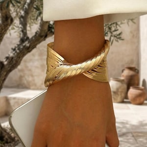 May include: A wide gold-tone cuff bracelet featuring a textured, multi-strand design that twists at the center. The metallic surface has a polished finish with fine, linear detailing across each individual strand, creating a layered appearance.