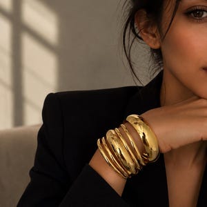 May include: A stack of five polished gold-tone metal bangles of varying thicknesses worn on the wrist. The bracelets feature a smooth, reflective surface and a rounded, curved profile, creating a layered appearance against a black blazer.