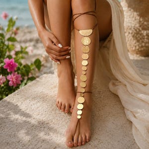 May include: A gold-toned leg chain featuring a series of circular metal discs connected in a vertical line, extending from a thigh strap down to a toe loop. The accessory is constructed with thin brown cord ties that wrap around the upper leg and ankle.
