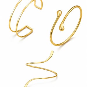 May include: Three gold-toned metal bracelets. One is a double-band open cuff, another is a thin wire bangle with a single teardrop-shaped terminal, and the third is a spiraling, wavy wire band that curves into a continuous, open-ended shape.
