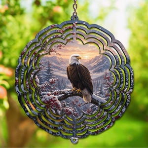 May include: A circular metal wind spinner featuring a scalloped edge and a printed image of a bald eagle perched on a snow-covered branch. The background depicts a winter mountain landscape at sunset with pine trees and red berries on the branches.