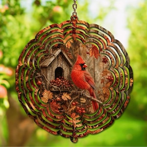May include: A circular metal wind spinner featuring a vibrant red cardinal perched on a branch next to a rustic wooden birdhouse, surrounded by autumn leaves and red berries, with a scalloped edge design and a silver hanging chain at the top.