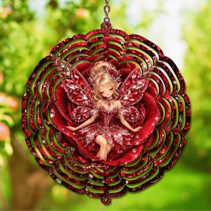 May include: A circular wind spinner featuring a central illustration of a fairy with blonde hair and red wings, surrounded by layers of deep red rose petals. The design incorporates a glittery finish and is suspended by a metal chain link.