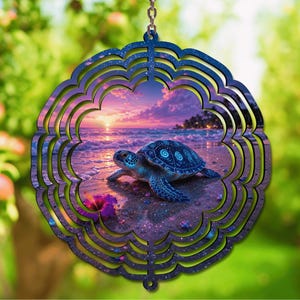 May include: A circular wind spinner featuring a scalloped edge and a dark blue frame. The center displays a sea turtle on a sandy beach at sunset, with purple and orange clouds, ocean waves, and a vibrant pink hibiscus flower in the foreground.