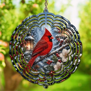 May include: A circular metal wind spinner featuring a vibrant red cardinal perched on a snowy branch. The design includes a glowing lantern, white flowers, and red berries against a winter forest background with a scalloped, layered edge.